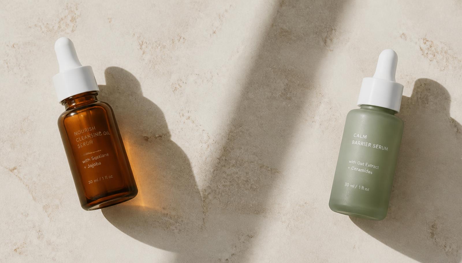 An amber serum dropper and a sage-green serum dropper placed apart on a cream stone surface in hard daylight