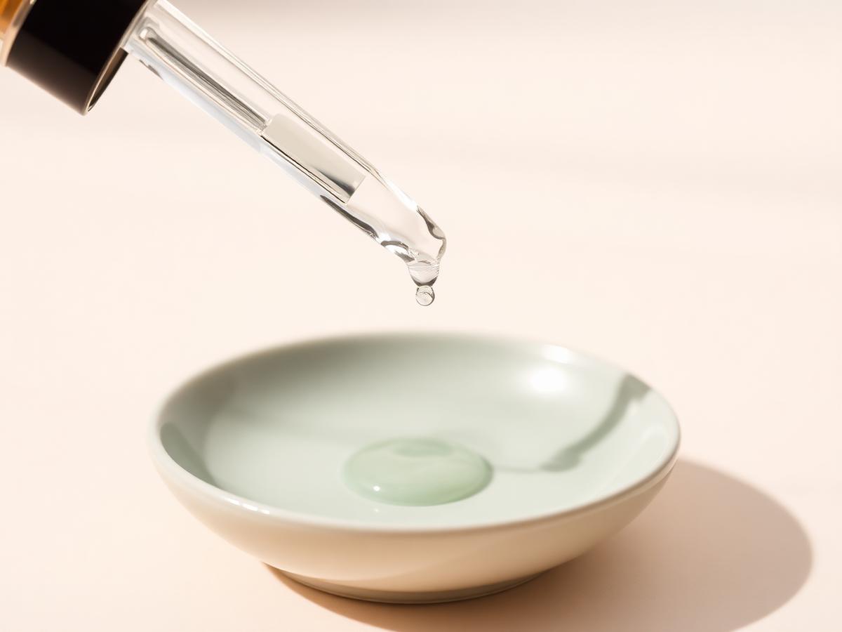 A glass dropper releasing a serum droplet into a pale sage ceramic dish